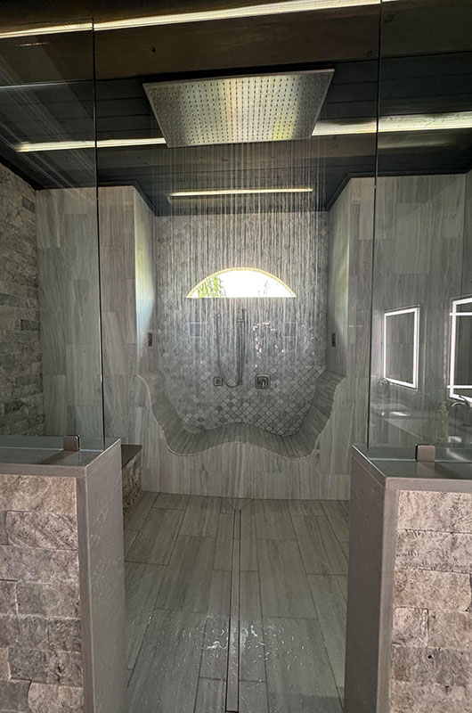 Luxury walk-in shower with stone walls, dual rainfall shower heads, a curved bench, large overhead rain shower, and natural light coming through a small arched window above the mosaic-tiled back wall.