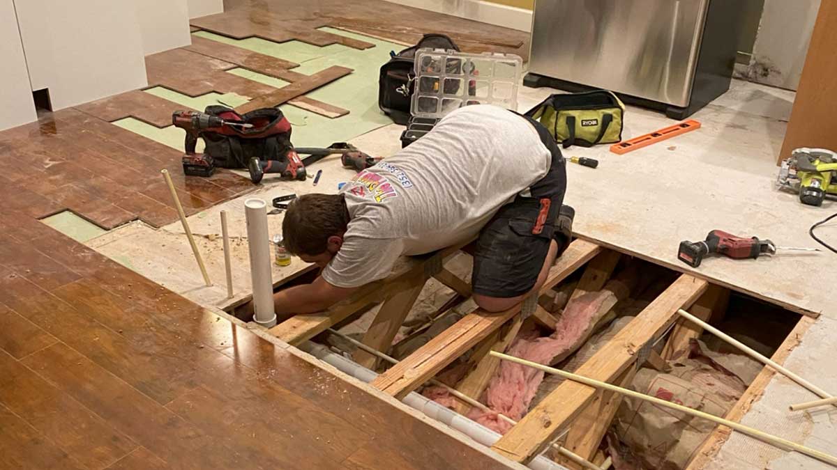 A person kneels on a partially dismantled wooden floor, working with exposed pipes and insulation. Tools and construction materials are scattered around the work area.