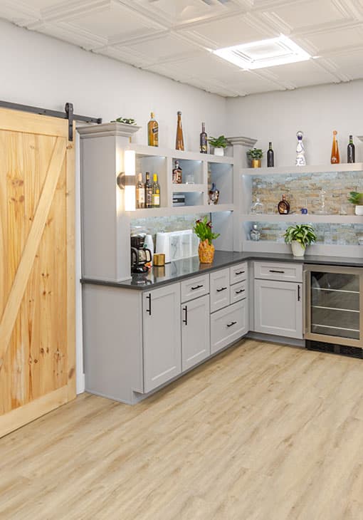 A modern kitchen with light gray cabinets, open shelves displaying bottles and decor, a black countertop, a wooden sliding barn door, potted plants, and light wood flooring.