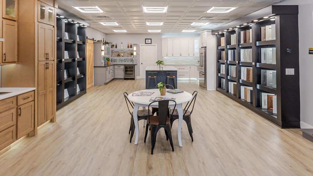 A modern showroom displays kitchen cabinets and countertops, with sample panels on dark shelves. A round table with four chairs sits in the center on light wood flooring, and a kitchen island is visible in the background.