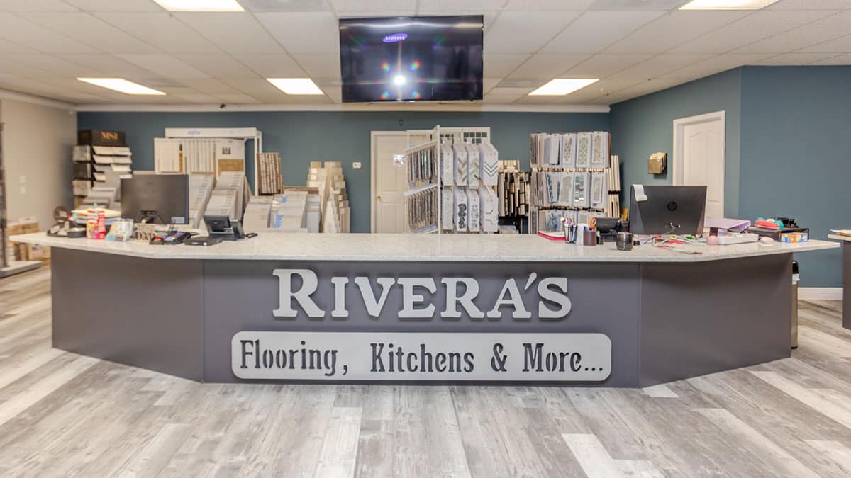 A wide view of a showroom reception desk with a sign reading "RIVERA'S Flooring, Kitchens & More..." showcasing flooring samples and kitchen design displays in the background.