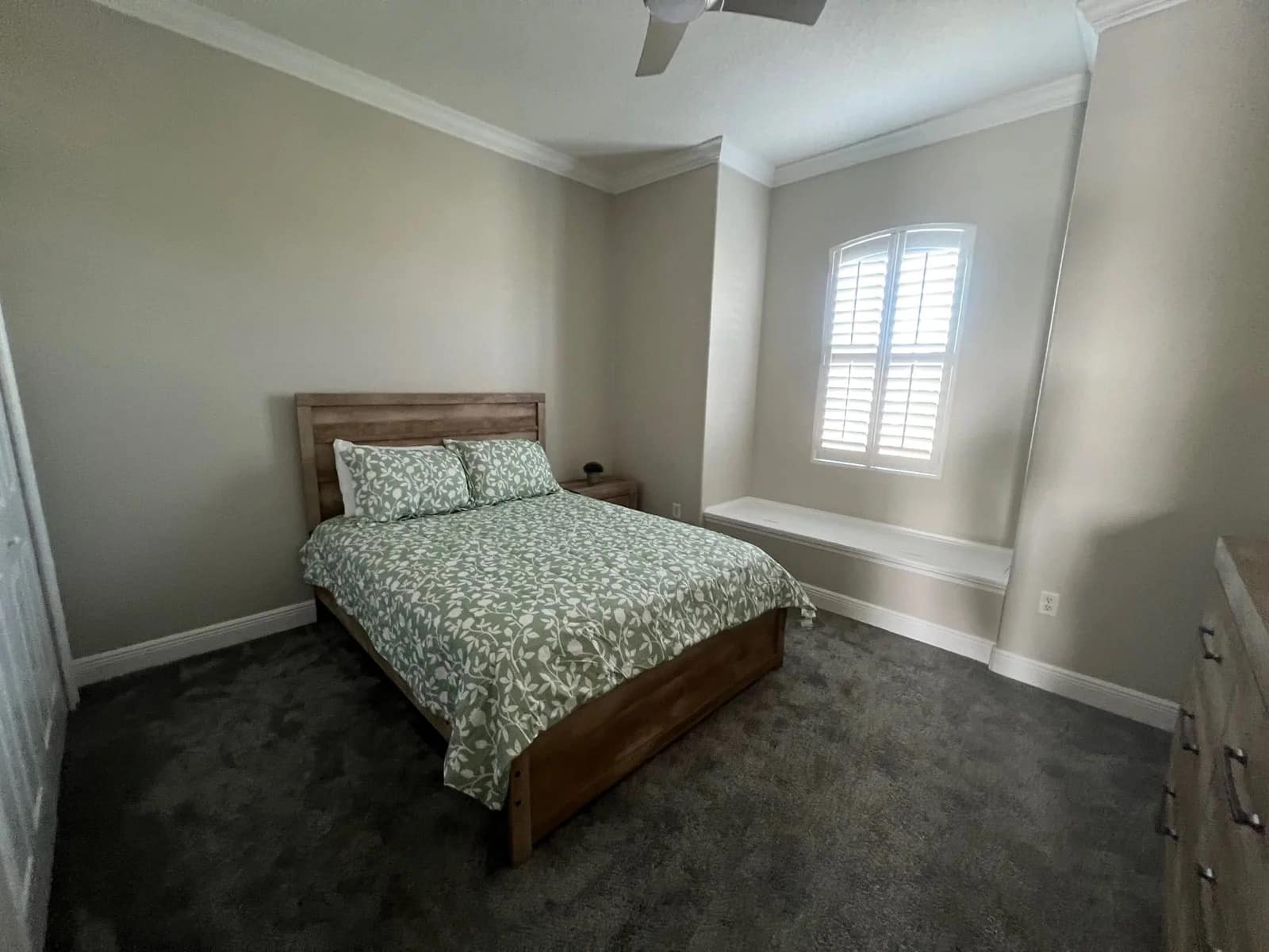 A bedroom with a wooden bed frame and green patterned bedding, gray carpet, light-colored walls, a ceiling fan, a chest of drawers, and a window with white shutters above a built-in bench.