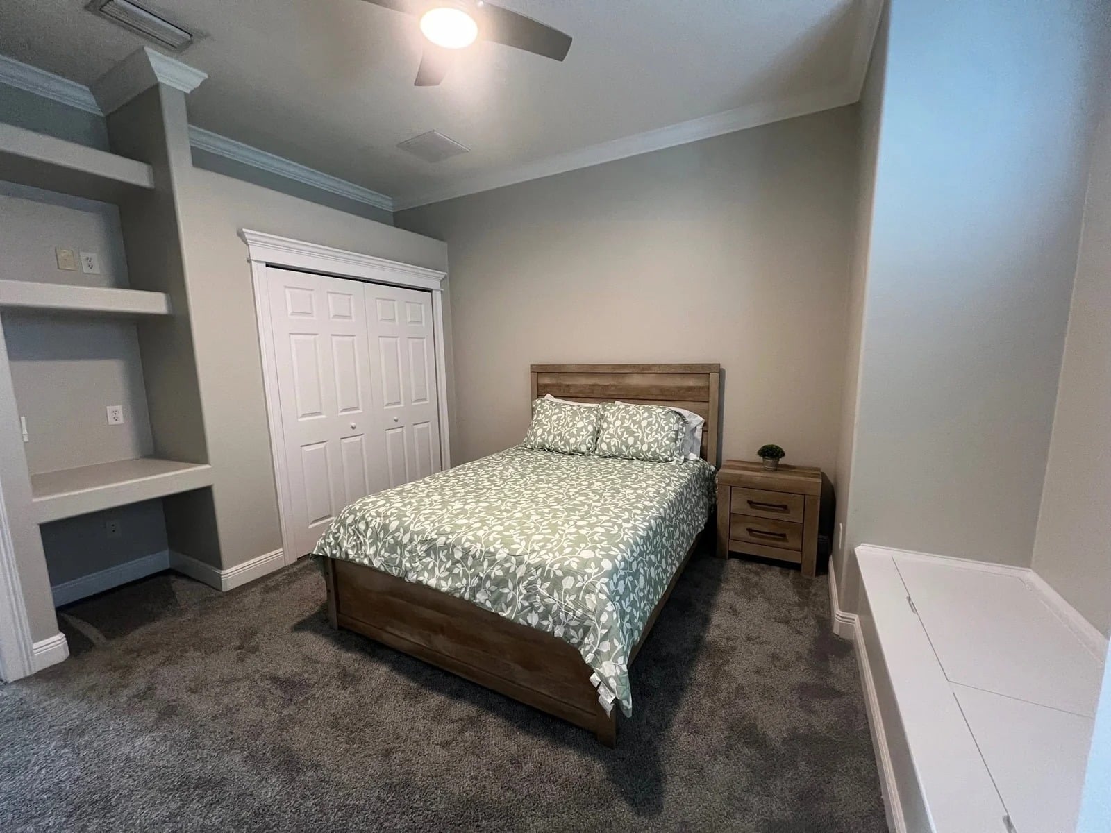 A neatly made bed with a green and white patterned comforter is centered in a carpeted bedroom. There’s a wooden nightstand, built-in shelves, a white closet, and a bench under a window. Walls are light gray.