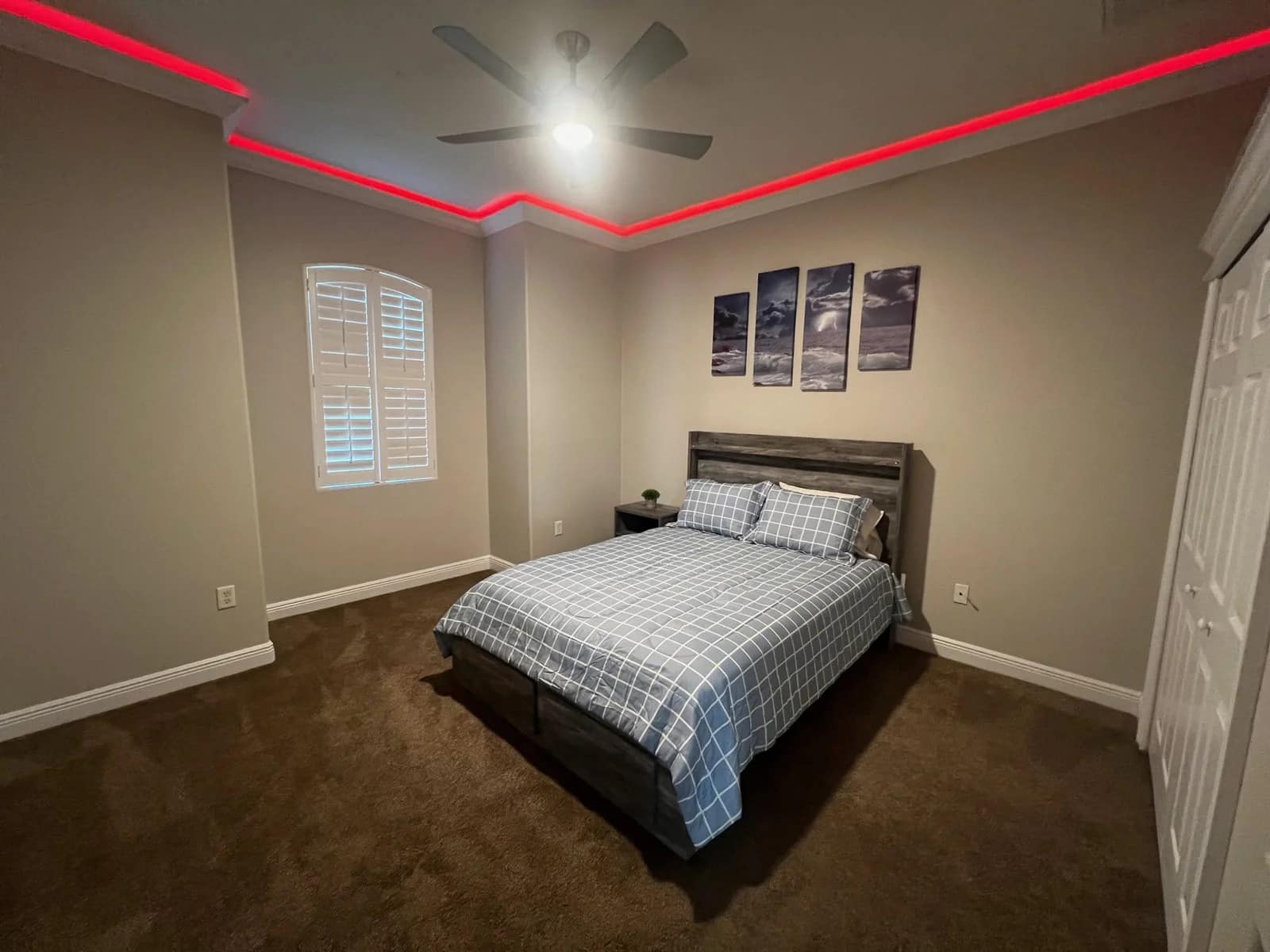 A bedroom with a neatly made bed with checkered bedding, a nightstand, three-panel wall art above the headboard, brown carpet, beige walls, a ceiling fan, and red LED strip lighting along the ceiling.