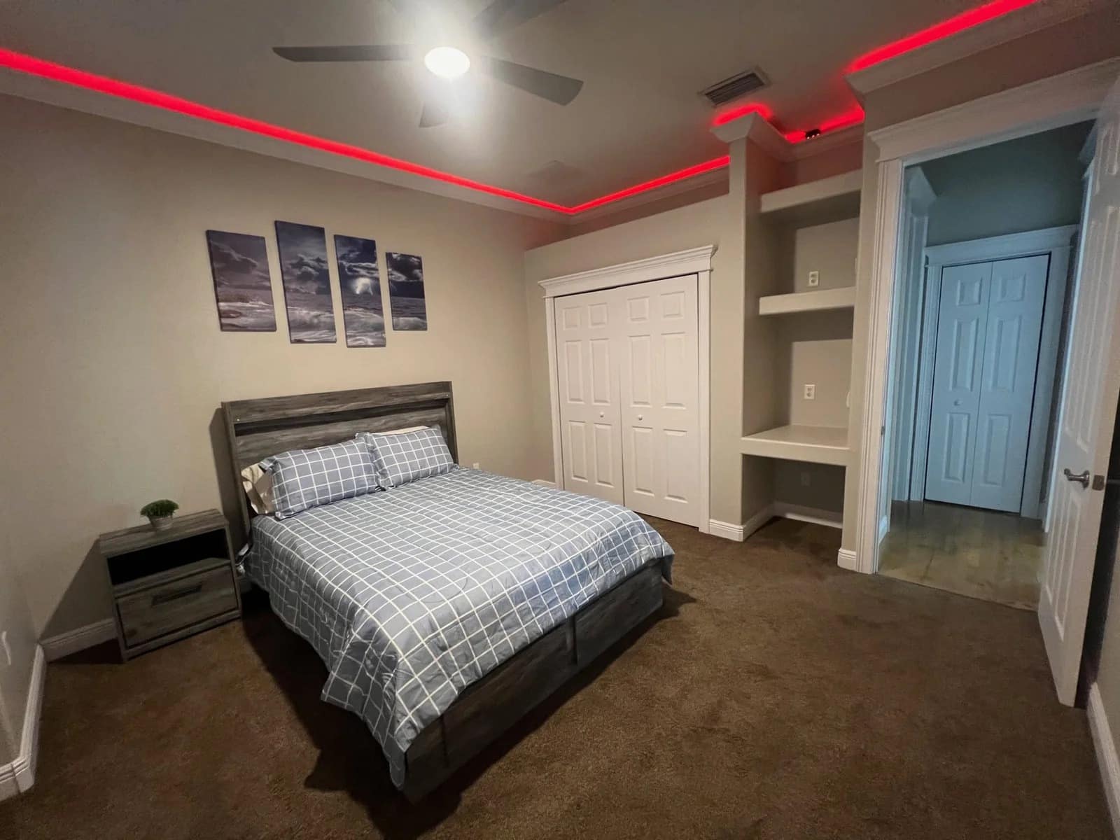 A modern bedroom with a bed, nightstand, closet doors, built-in shelves, brown carpet, light walls, and red LED strip lights along the ceiling. Three landscape pictures hang above the bed.