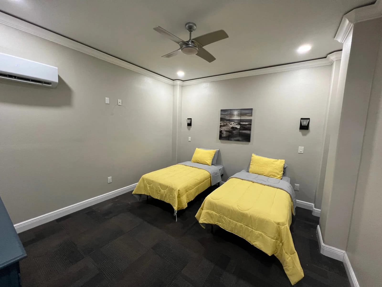 A minimalist bedroom with two single beds featuring yellow bedding, a ceiling fan, a wall-mounted air conditioner, and a seascape picture above the beds. The walls are light-colored and the floor is dark.