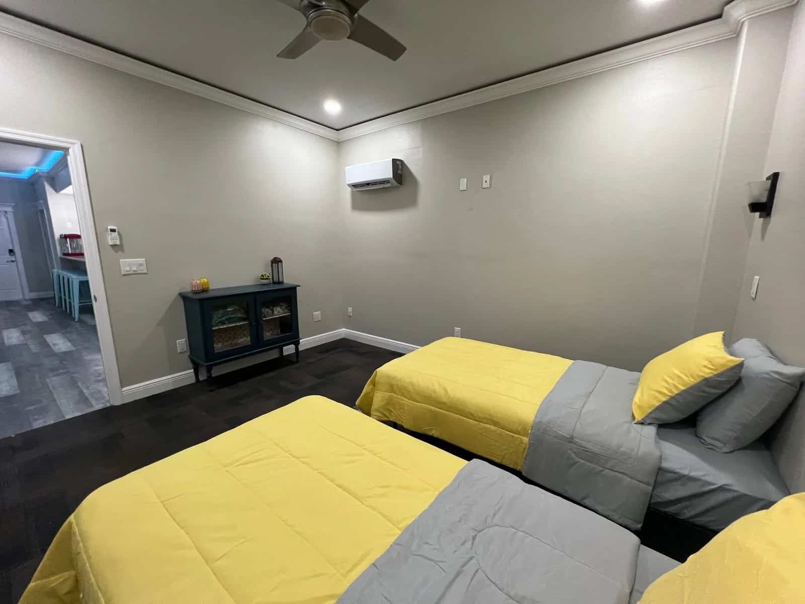 A bedroom with two twin beds featuring yellow and gray bedding, a small cabinet with glass doors, wall-mounted air conditioning, and a ceiling fan. An adjacent room with blue lighting is visible through an open doorway.