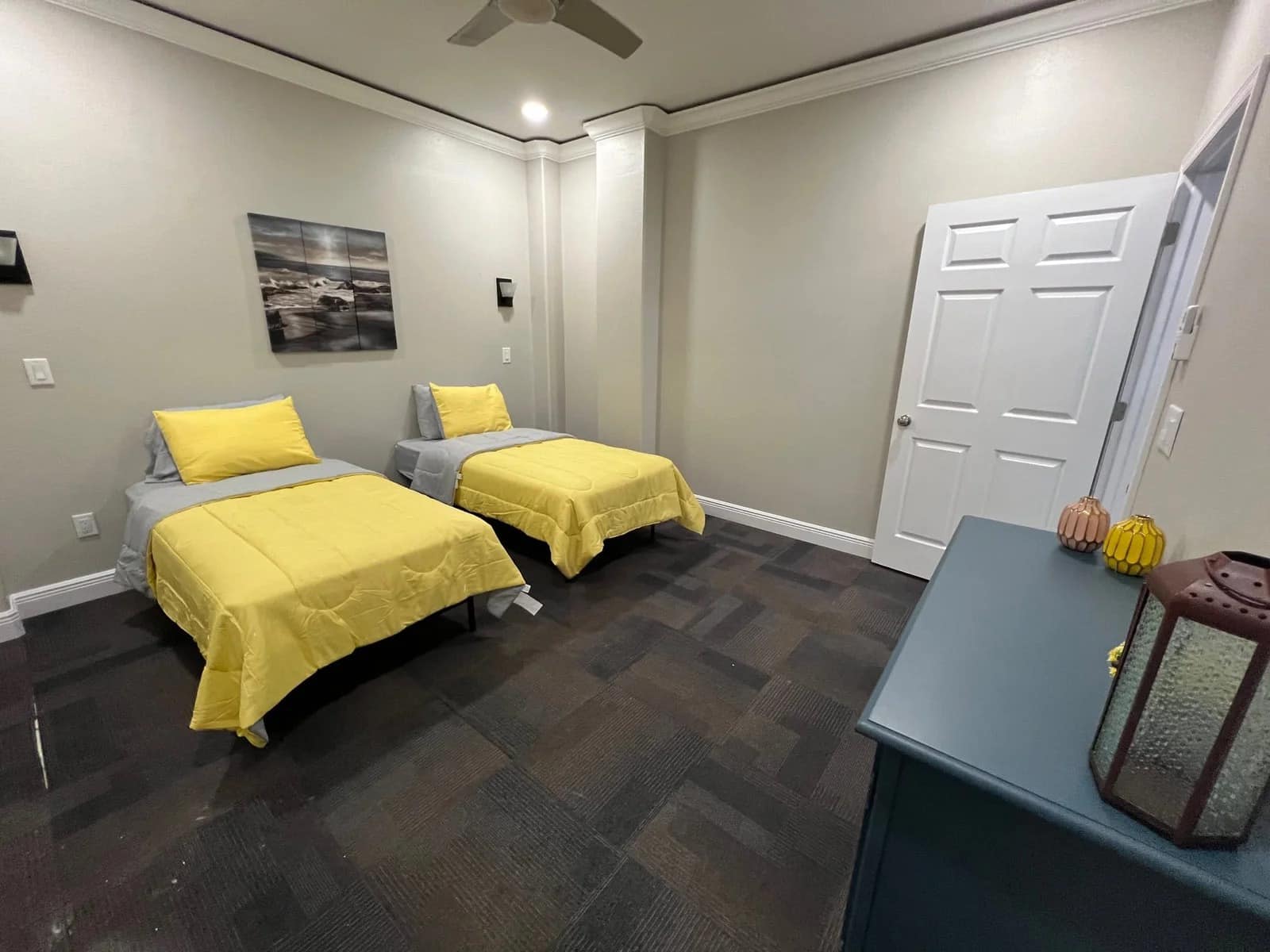 A clean bedroom with two single beds, each with yellow bedding, set against light-colored walls. A dresser with decor and a lantern sits near a white door. A seascape photo hangs above one bed.