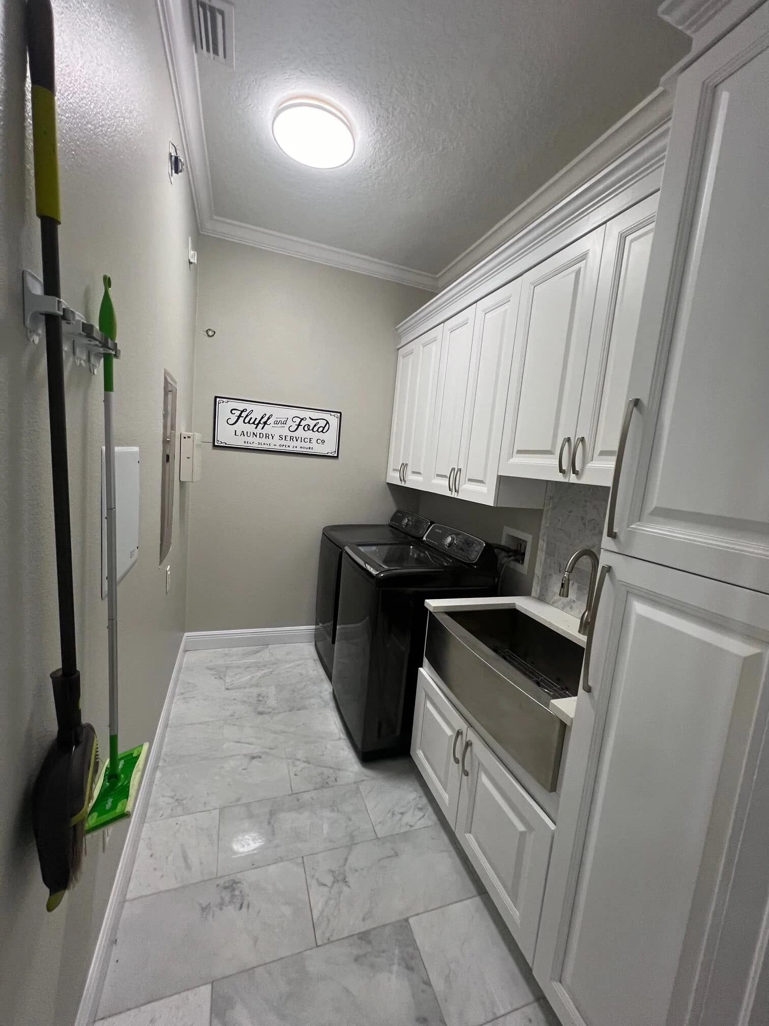 A modern laundry room with white cabinets, a black washer and dryer, a utility sink, marble tile floor, and cleaning tools mounted on the left wall. A decorative sign is hung above the appliances.