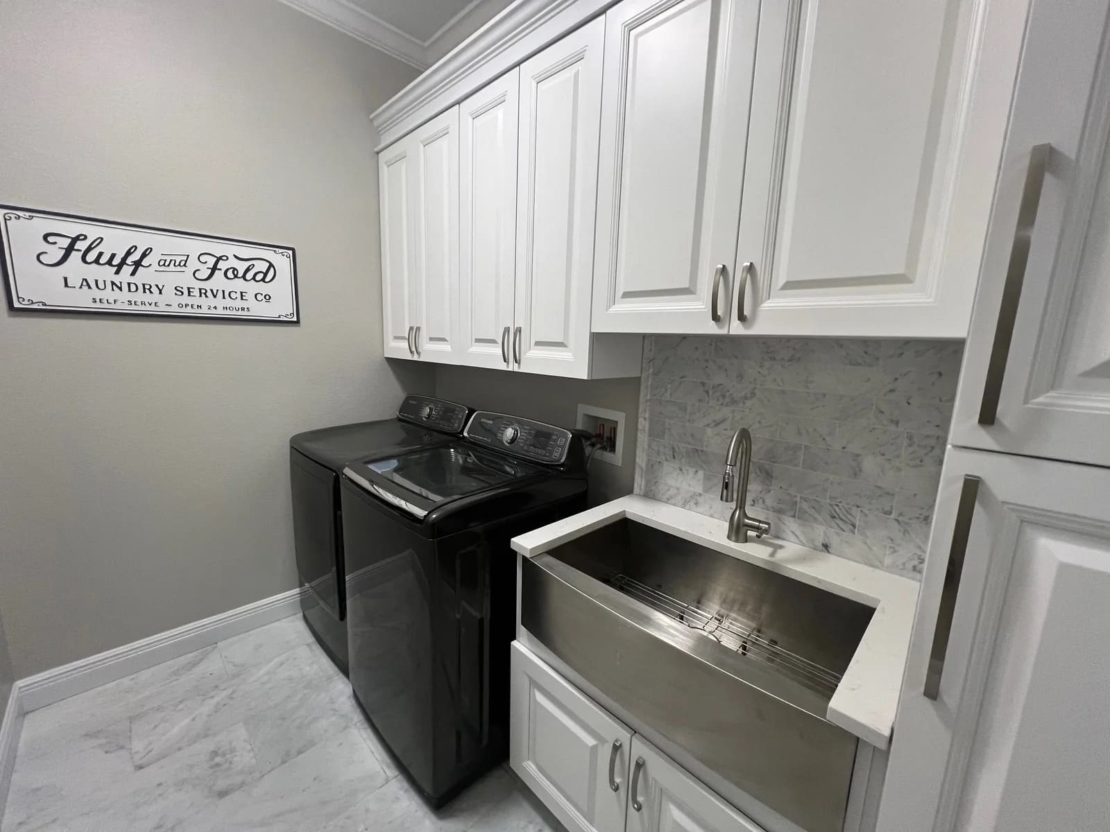 A modern laundry room with white cabinets, a stainless steel utility sink, a black washer and dryer, marble tile floor, and a wall sign that reads "Fluff and Fold Laundry Service Co.
