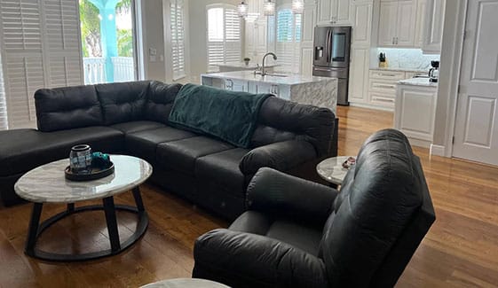 A modern living room with black leather sofas and armchairs, two round marble coffee tables, hardwood floors, and a white kitchen with stainless steel appliances in the background. Sunlight streams in through large windows.