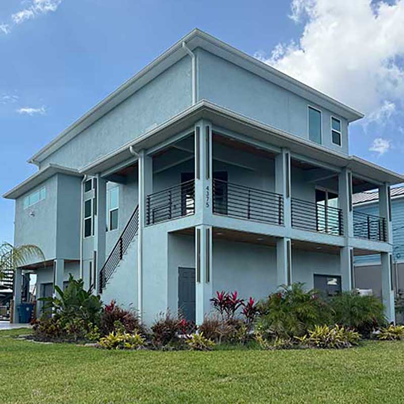A modern two-story light blue house with a large wraparound balcony, raised on stilts, surrounded by tropical plants and a green lawn under a partly cloudy sky.