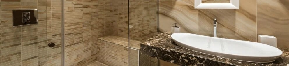 Modern bathroom with beige and brown tiled walls, a glass-enclosed shower with a built-in bench, and a black marble countertop with a white oval sink and a rectangular mirror above it.