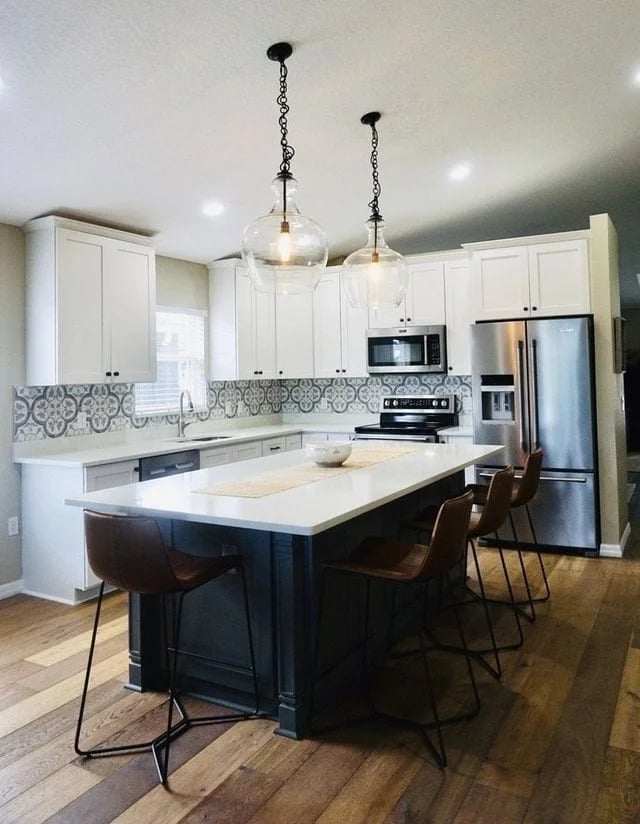Modern kitchen with white cabinets, patterned tile backsplash, stainless steel appliances, wood flooring, and a large island with four brown barstools. Two glass pendant lights hang above the island.