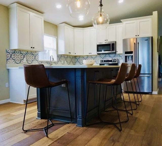 A modern kitchen with white cabinets, blue patterned tile backsplash, stainless steel appliances, a large blue island, and four brown barstools with metal legs on light wood flooring.