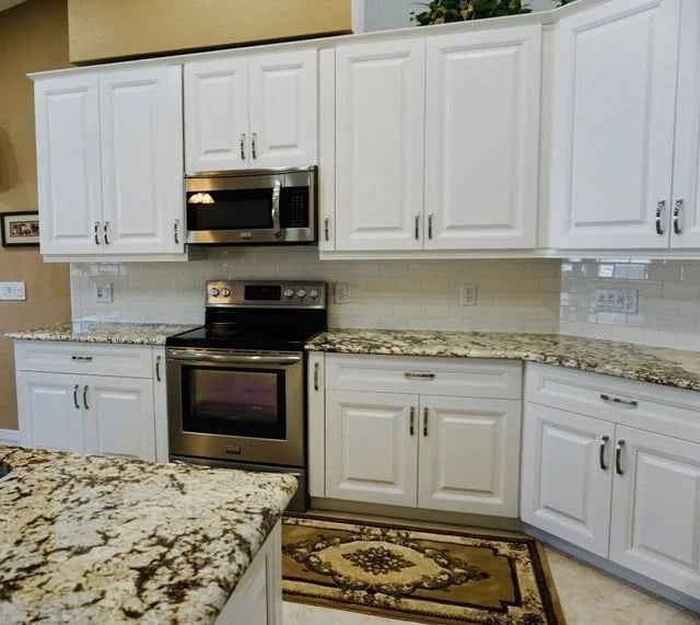A modern kitchen with white cabinets, granite countertops, stainless steel appliances including a stove and microwave, white subway tile backsplash, and a decorative rug on the floor.