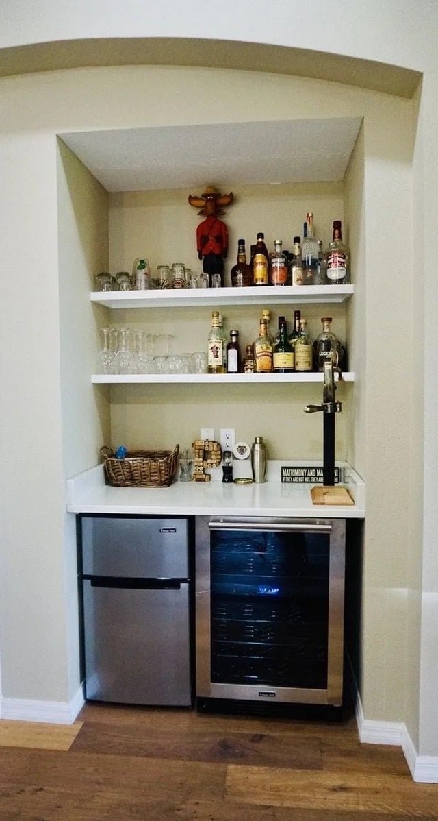 A small home bar set in a wall alcove with glass shelves holding bottles, glasses, and decor. Below are a mini fridge and wine cooler, with a countertop featuring a basket, cocktail tools, and a beer tap.