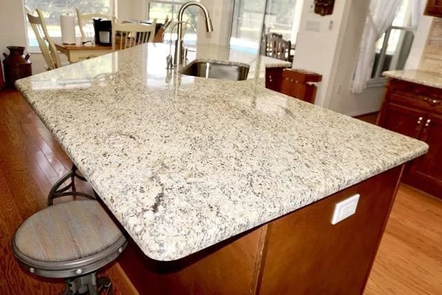 A polished granite kitchen island with rounded edges, brown wooden base, and a single bar stool. Sink and faucet are visible, with hardwood flooring and natural light from windows in the background.