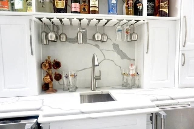A small home bar sink with white cabinets, a marble-look backsplash, hanging wine glasses, assorted liquor bottles above, and various glasses and a wooden mug holder on the counter.