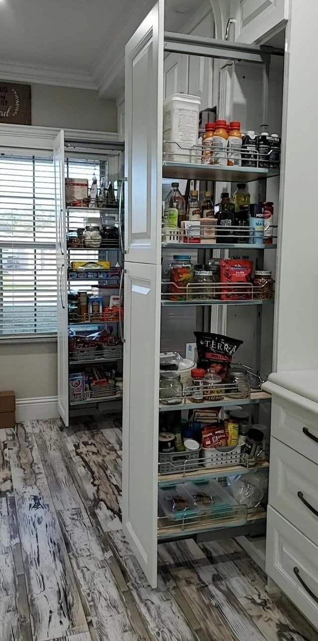 A modern kitchen pantry with tall white pull-out cabinets neatly organized with jars, cans, snacks, and kitchen supplies. Sunlight streams in through a window with blinds, illuminating the wood-patterned floor.