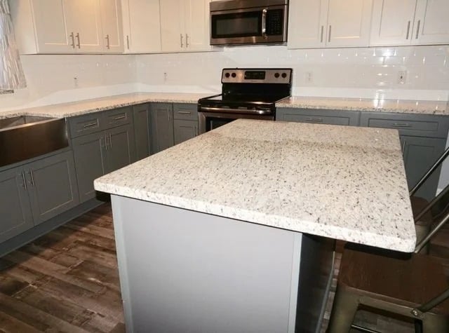 Modern kitchen with white and gray cabinets, stainless steel appliances, a large kitchen island with a speckled granite countertop, and wooden flooring. A brown chair is partially visible near the island.