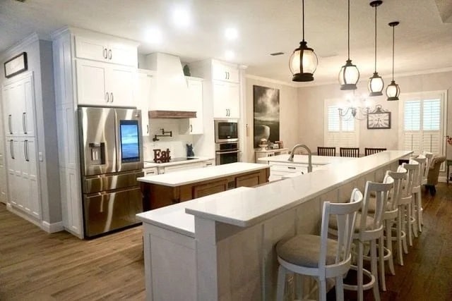 A spacious modern kitchen with white cabinets, stainless steel appliances, a large island with a white countertop, bar stools, pendant lights, and hardwood floors. Dining table and wall art are visible in the background.