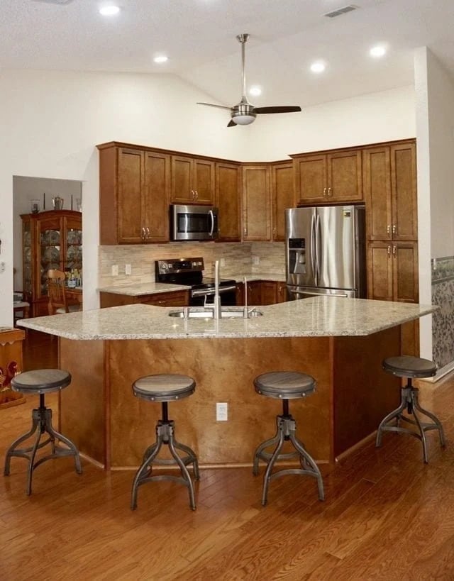 Modern kitchen with wooden cabinets, stainless steel appliances, and a large granite island featuring a sink and four metal bar stools. Hardwood floors and neutral walls complete the space. Ceiling fan overhead.