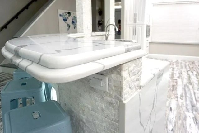 A marble kitchen counter with rounded edges, white stone backsplash, and light blue bar stools. The floor is white with gray streaks, and a world map decor is visible on the wall in the background.