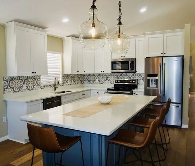 Modern kitchen with white cabinets, stainless steel appliances, patterned tile backsplash, and a large island with a white countertop. Four brown chairs line one side of the island, and two glass pendant lights hang above.