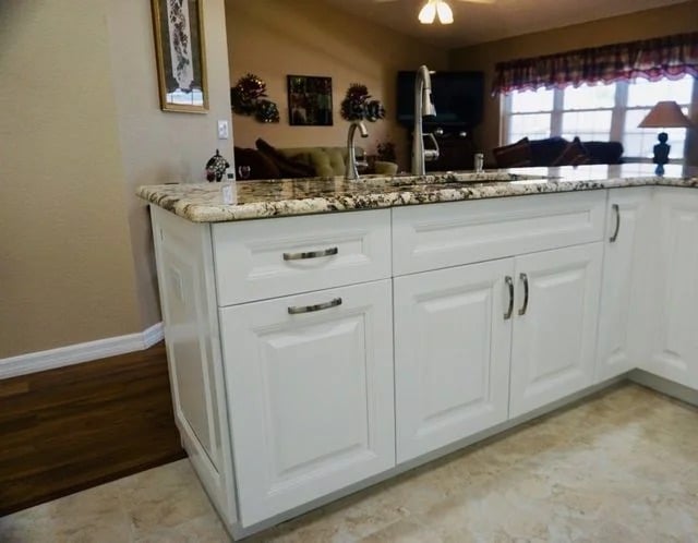 White kitchen island with granite countertop, silver handles, and a built-in sink. The background shows a living area with a TV, windows, and wall decorations. The floor is light tile.