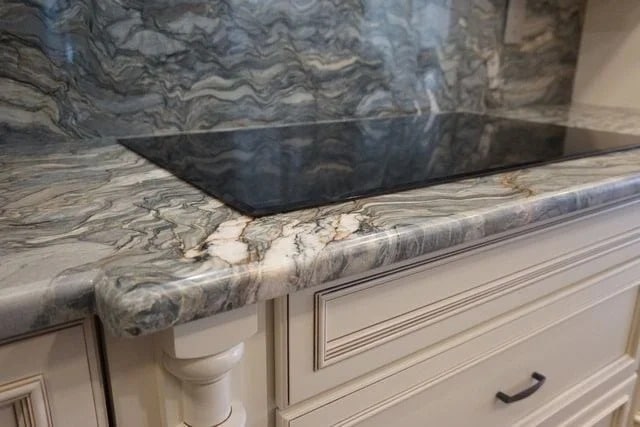 A close-up of a kitchen countertop with a marble-like surface featuring swirling gray, white, and beige patterns, an electric cooktop, and cream-colored drawers beneath.