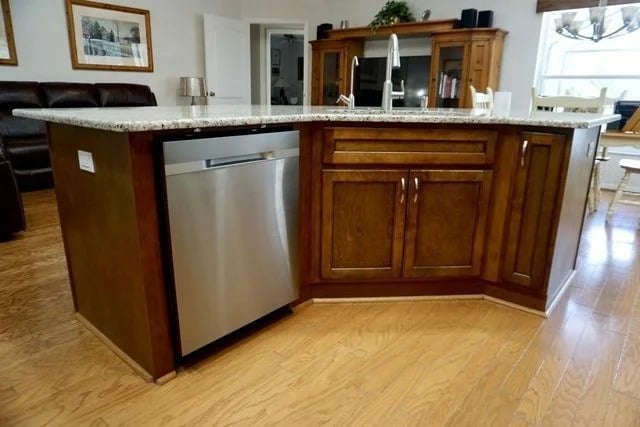 A kitchen island with brown wooden cabinets, a built-in stainless steel dishwasher, and a granite countertop, set on a light wood floor in a bright, open living space.