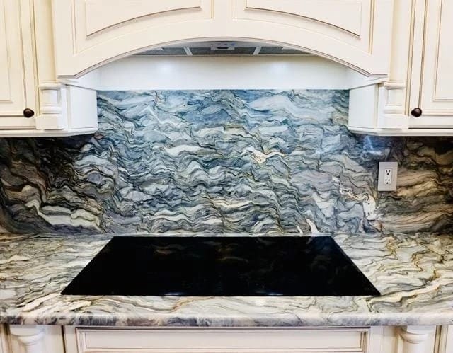 A kitchen countertop and backsplash made of marble with swirling gray, white, and blue patterns, featuring a black glass cooktop and off-white cabinetry above and on the sides.
