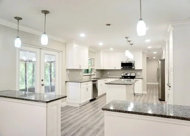 Modern kitchen with white cabinets, gray granite countertops, stainless steel appliances, pendant lights, an island in the center, light wood flooring, and French doors leading outside.