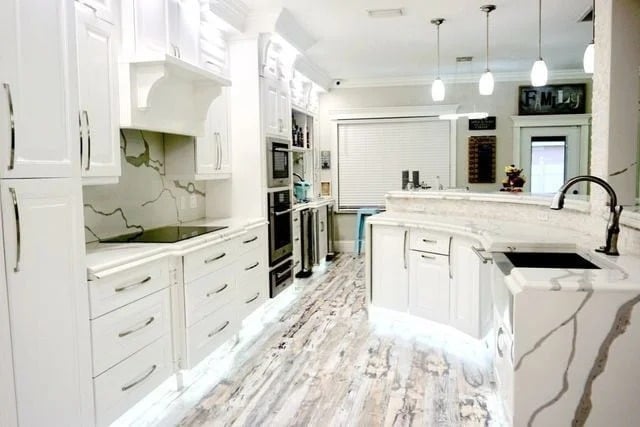 Modern kitchen with white cabinets, marble countertops, and backsplash. Pendant lights hang over a curved island with a sink; the floor features a whitewashed wood pattern. The space is bright and open with sleek appliances.