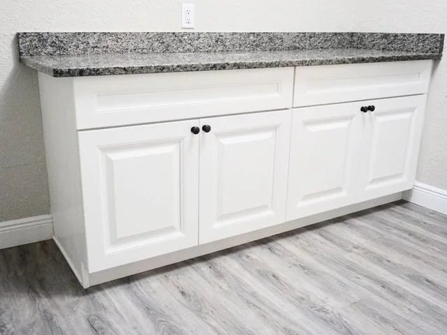 White kitchen cabinet with two drawers and four doors, topped with a gray granite countertop, set against a light-colored wall and gray wood-look flooring.