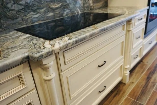 A kitchen countertop with a swirling gray and white marble pattern, cream-colored cabinets with black handles, and a built-in black glass cooktop are shown. The floor is wood with a brown finish.