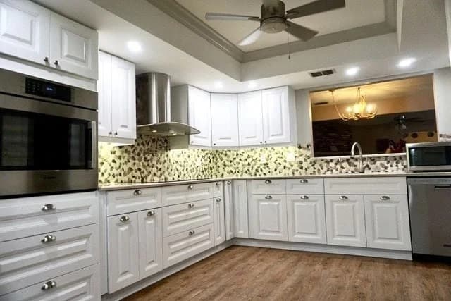 Modern kitchen with white cabinets, stainless steel appliances, mosaic tile backsplash, wooden floor, a ceiling fan, and recessed lighting. A window above the sink reveals a chandelier in the adjacent room.