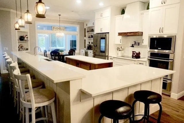 Modern kitchen with white cabinets, a large island, stainless steel appliances, pendant lights, bar stools, and a dining area with large windows in the background. Warm, inviting atmosphere.