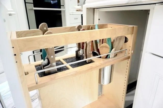 A wooden pull-out kitchen drawer organizer holds knives, cooking utensils, and wooden spoons, with a metal cup attached on the side, set in a white kitchen cabinet.