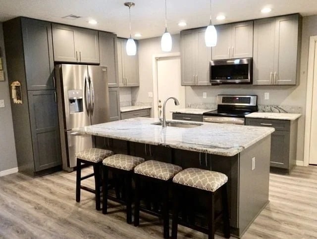 Modern kitchen with gray cabinets, stainless steel appliances, marble countertops, and an island with a sink. Four patterned stools are placed under the island, and pendant lights hang overhead. Wood-style flooring completes the space.