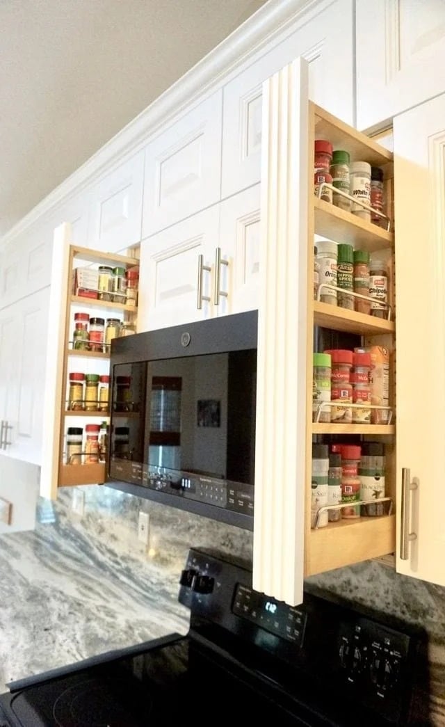 Two pull-out spice racks are built into kitchen cabinets on either side of a black microwave above a stove, displaying neatly organized spice jars on shelves. The cabinets and kitchen decor are white.
