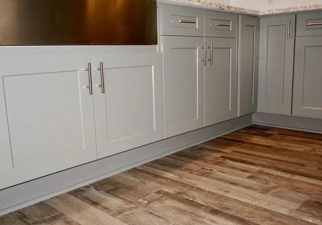 Light gray kitchen cabinets with silver handles are set against a light-colored granite countertop. The floor is covered with wood-style vinyl planks in shades of brown and gray.