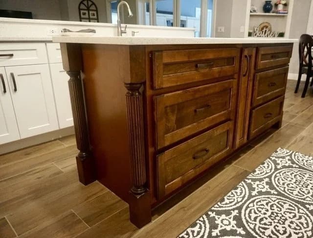 A wooden kitchen island with ornate legs and multiple drawers stands on a wood floor next to a white countertop and white cabinets, with a patterned rug partially visible in the foreground.