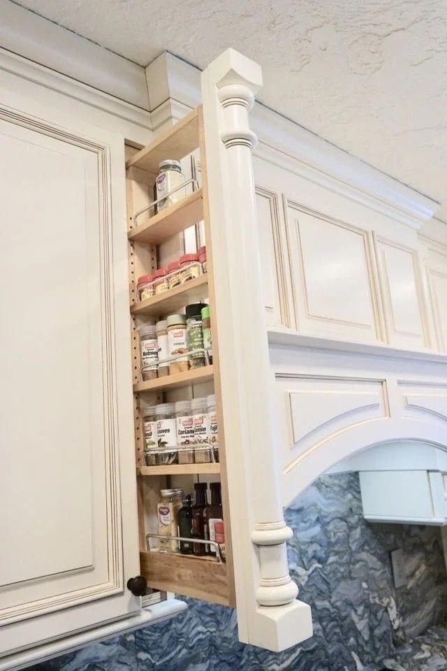 A tall, narrow pull-out spice rack is built into a white kitchen cabinet, neatly holding various spice jars and bottles. The rack is extended outward, revealing multiple organized shelves.