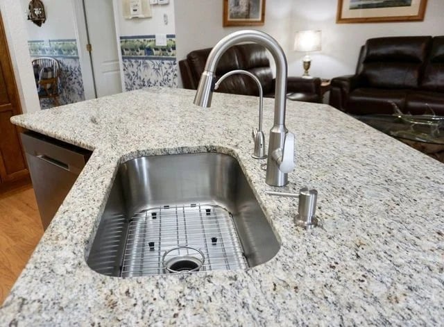 A modern stainless steel kitchen sink with a high-arc faucet is set in a granite countertop on an island, with a living room visible in the background.