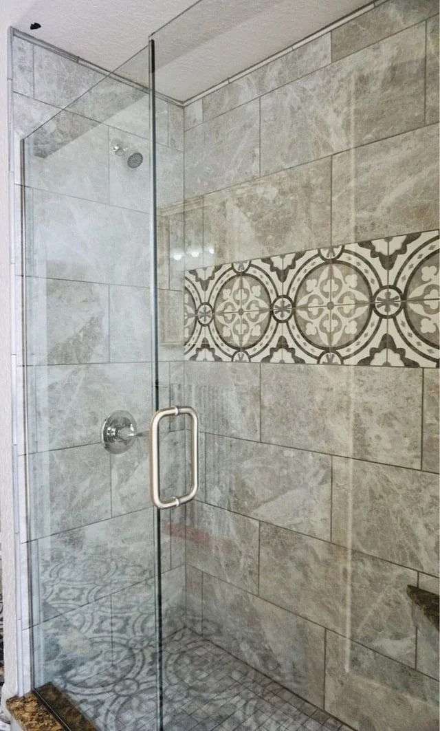 A modern shower with gray marble tiles, a glass door, and a decorative horizontal strip of patterned tiles featuring circular and geometric designs. The floor consists of small square tiles in coordinating tones.