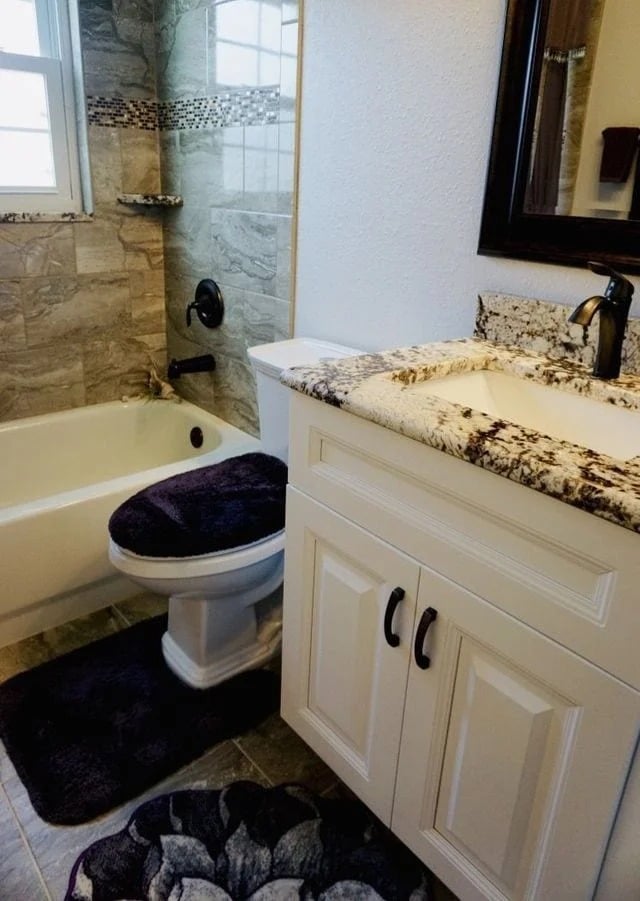 A small bathroom with a bathtub-shower combo, a toilet with a dark purple cover, a matching bath mat, and a white vanity with a granite countertop and a black faucet. Light gray tile and mosaic accents in the shower.