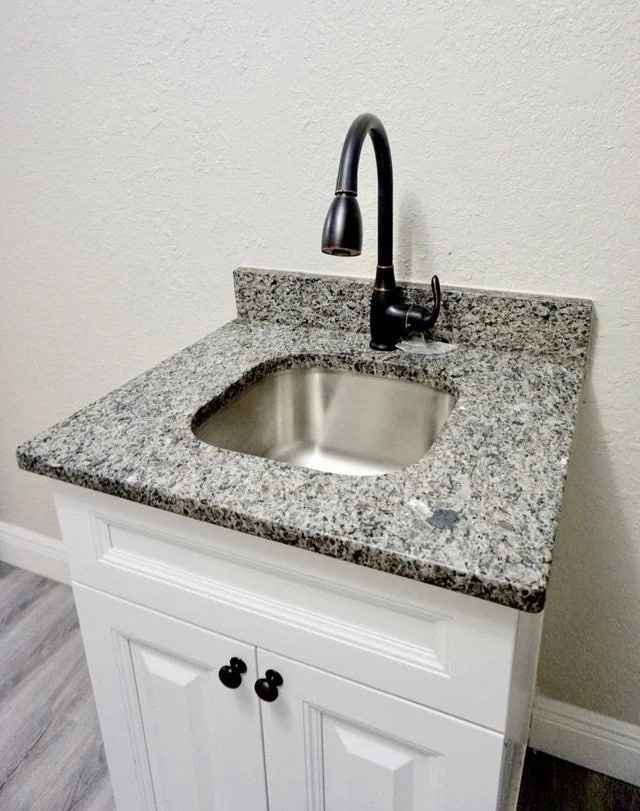 A small sink with a granite countertop, modern black faucet, and white cabinet with double doors, set against a light-colored textured wall and gray wood-look flooring.