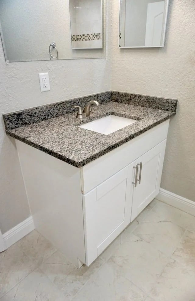 A bathroom vanity with a granite countertop, white sink, and white cabinet with silver handles, set against a beige wall with mirrors above and light-colored tile flooring.